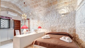 Een slaapkamer met een stenen muur, een bed met een bruine sprei, een witte nachtkastje met een rode lamp, en een klein wit tafeltje met een decoratief object.