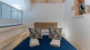 Une chambre avec un lit, deux oreillers, une fenêtre avec des stores, une tête de lit en bois et une petite étagère sur laquelle est posé un objet décoratif.
