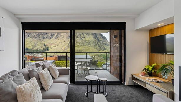 A modern living room with a sofa, a coffee table, and a TV. It features a balcony with a mountain view. - Kiwi Studio Apartment minutes from Coronet Peak (Queenstown)