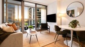 Una sala de estar moderna con un sofá, una mesita con libros, un área de comedor con sillas, un televisor montado en la pared y un amplio ventanal con vistas a la ciudad.