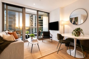 A modern living room with a sofa, a small table with books, a dining area with chairs, a TV mounted on the wall, and a large window with a city view.