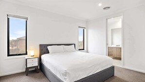A bedroom with a large bed, a nightstand, a window, and a bathroom visible through an open door. - Spacious 5-Bed Home in Peaceful Point Cook (Point Cook)