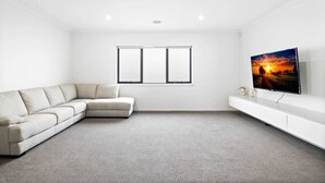 A modern living room with a beige sectional sofa, a flat-screen TV on a white entertainment unit, and a large window with blinds. - Spacious 5-Bed Home in Peaceful Point Cook (Point Cook)