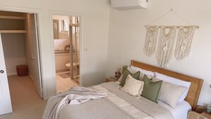 Une chambre à coucher avec un lit, une table de chevet, une décoration murale en macramé et une porte ouverte donnant sur une salle de bain.
