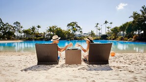 Twee personen zitten op rieten stoelen bij een zwembad aan het strand, met helderblauw water en palmbomen op de achtergrond.