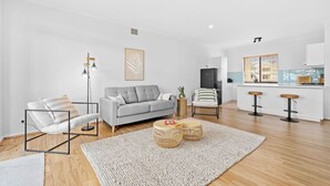 Un salón moderno con un sofá gris, un sillón blanco y una mesa de centro. Hay una zona de cocina con un refrigerador y taburetes de bar.