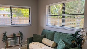 Ein Wohnzimmer mit einem grünen Sofa, einem runden Holztisch und einem kleinen Beistelltisch. Zwei Fenster mit Jalousien, eine Heizung und ein Blumentopf sind vorhanden.