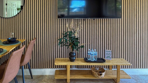 Ein moderner Essbereich mit einer Holzvertäfelung, einem runden Spiegel, einem an der Wand montierten Fernseher, einem Holztisch mit Stühlen, einer Vase mit Pflanzen und einem Willkommensschild.