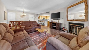 Ein geräumiges Wohnzimmer mit einer großen Eckcouch, einem Couchtisch und einem Kamin. An der Wand ist ein Fernseher angebracht, und im Hintergrund befindet sich eine Küchenzeile.