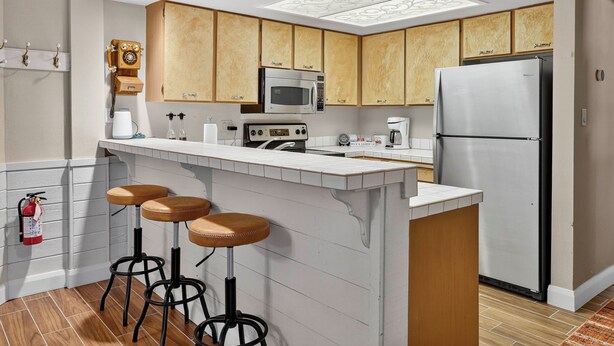 Eine moderne Küche mit Holzausstattung, einem Edelstahlkühlschrank, einer Mikrowelle und einer Kaffeemaschine. Drei Hocker stehen an der Bar.
