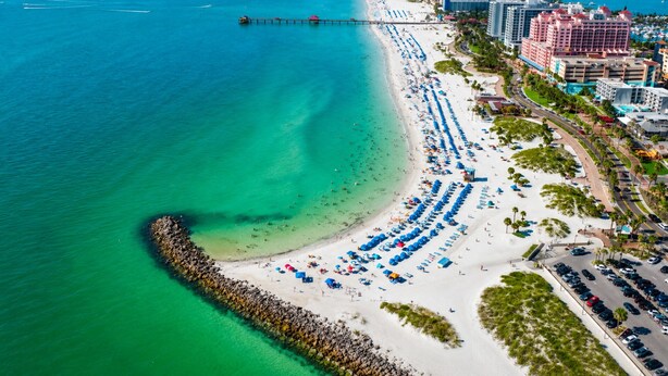 An aerial view of a beach with a pier extending into the ocean, lined with beach umbrellas and a row of hotels. - Island Vibes - 3BR Pool Home near St. Pete Beach (St. Petersburg)