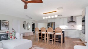 A modern kitchen with white cabinets, a central island, and a dining area with wooden chairs. - Island Vibes - 3BR Pool Home near St. Pete Beach (St. Petersburg)