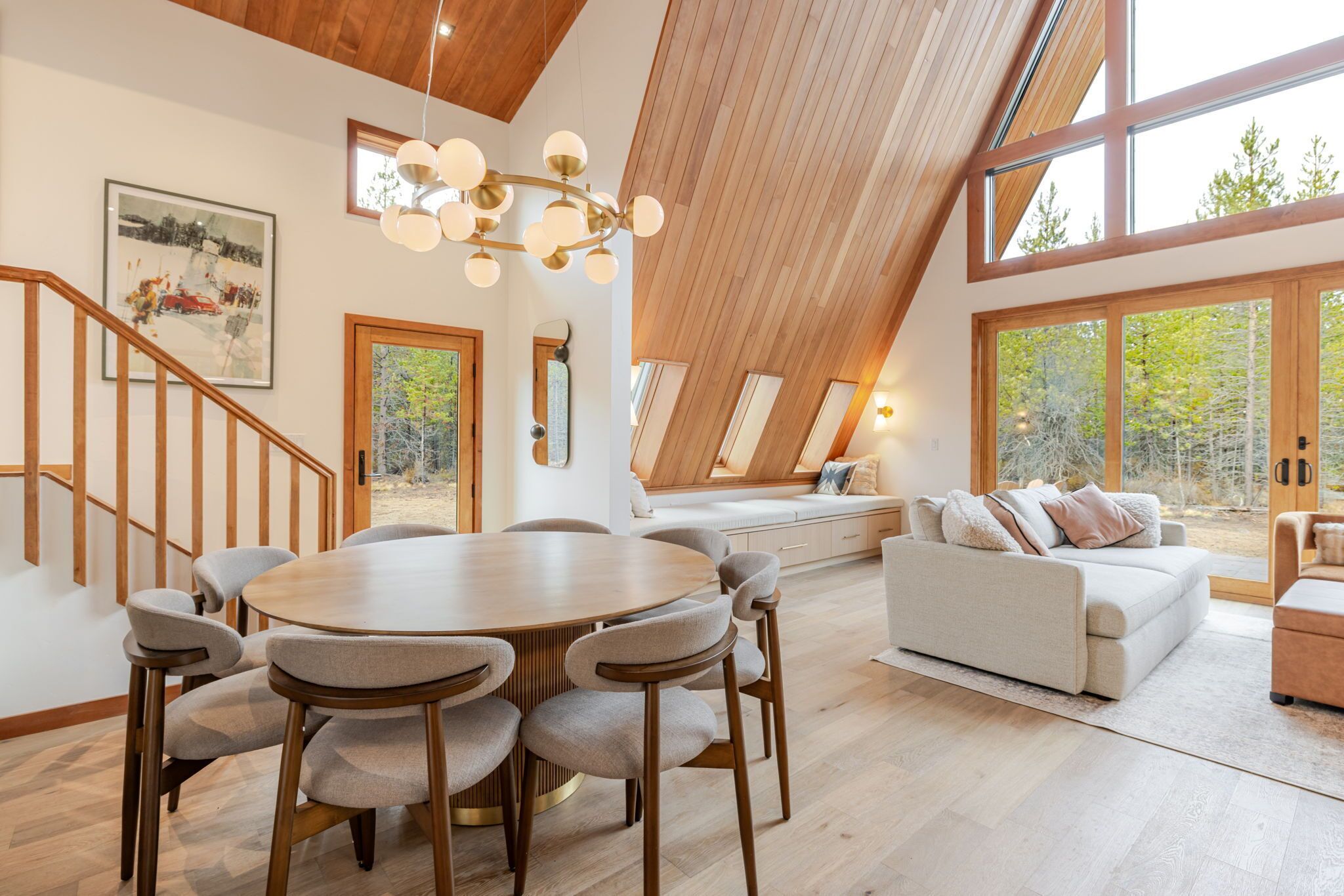 A spacious loft with a high ceiling, a dining area with a round table and chairs, a sofa, and large windows offering a view of the outdoors.