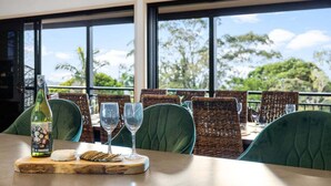 Una mesa de comedor con una botella de vino, queso, galletas y copas, con vistas a árboles y al cielo a través de amplios ventanales.