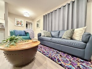 Un salon avec un canapé bleu, une table basse ronde, un tapis coloré et un rideau gris.