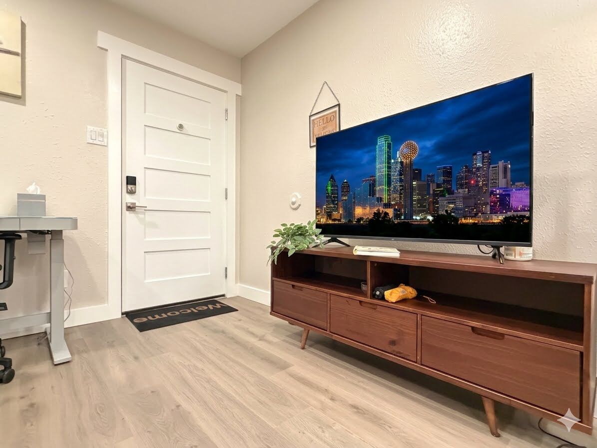 Een woonkamer met een flatscreen televisie, bevestigd aan een houten dressoir, een deur en een klein bureau met stoel.