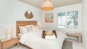 Quarto com cama de madeira, roupa de cama branca, cabeceira de tecido, luminária de mesa, tapeçaria na parede, janela com persianas e um banco junto à janela.