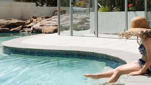 Uma piscina com água cristalina, borda de pedra e uma pessoa sentada à beira da piscina.