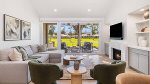 A modern living room with a large sofa, two armchairs, a coffee table, and a fireplace. There is a TV mounted above the fireplace and a sliding glass door leading to a patio with a view of a golf course. - Palm Desert Golf Course Condo w/ Patio Views, Fireplace, Resort Pool & Mountain Backdrop (Palm Desert)