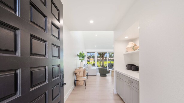 A modern interior with a dark wooden door, a seating area with a sofa and chairs, a kitchen with white cabinets, and a view of a green outdoor area through large windows. - Palm Desert Golf Course Condo w/ Patio Views, Fireplace, Resort Pool & Mountain Backdrop (Palm Desert)