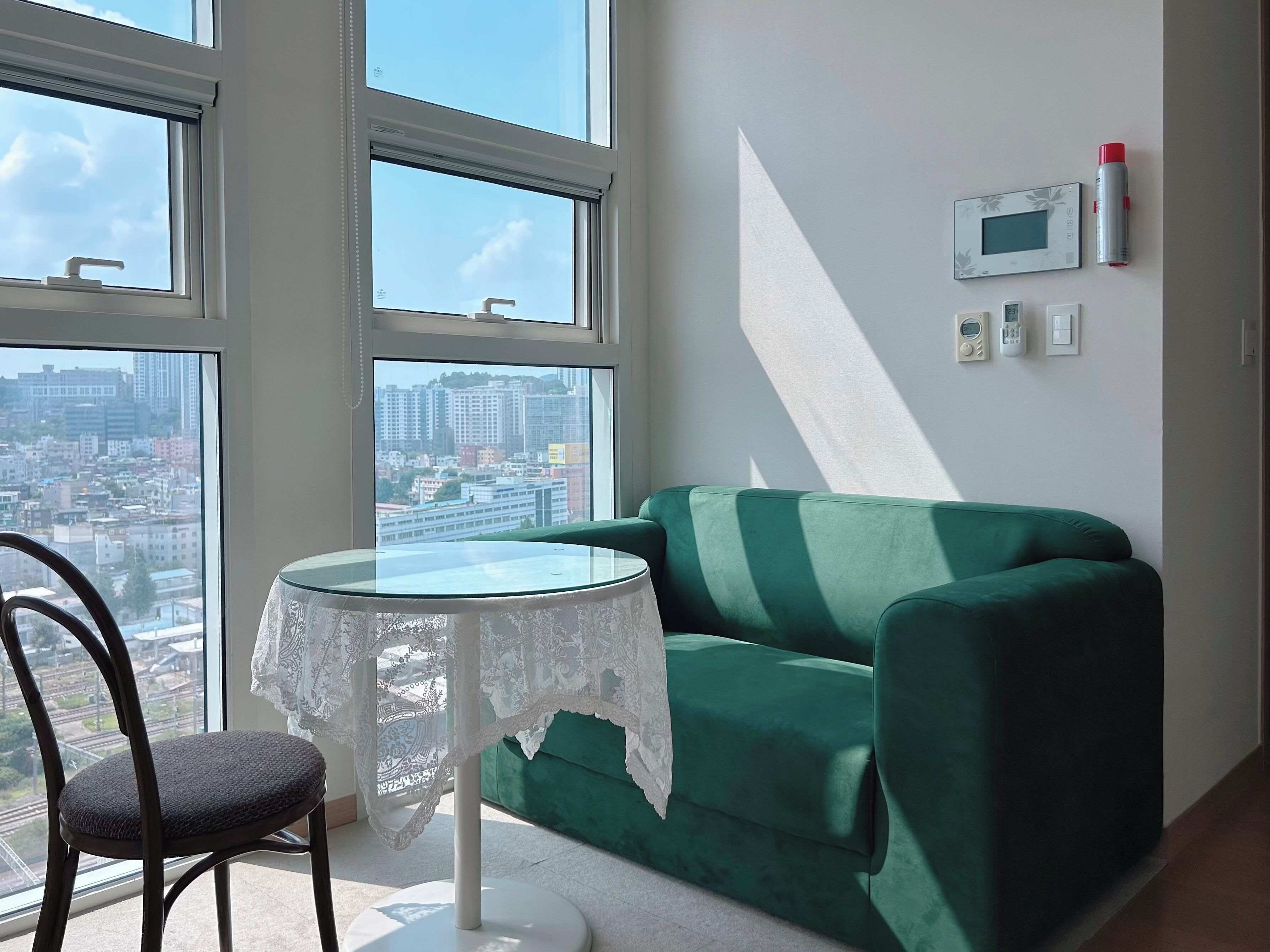 A room with a green sofa, a round table with a white tablecloth, a chair, and a view of a cityscape through large windows.