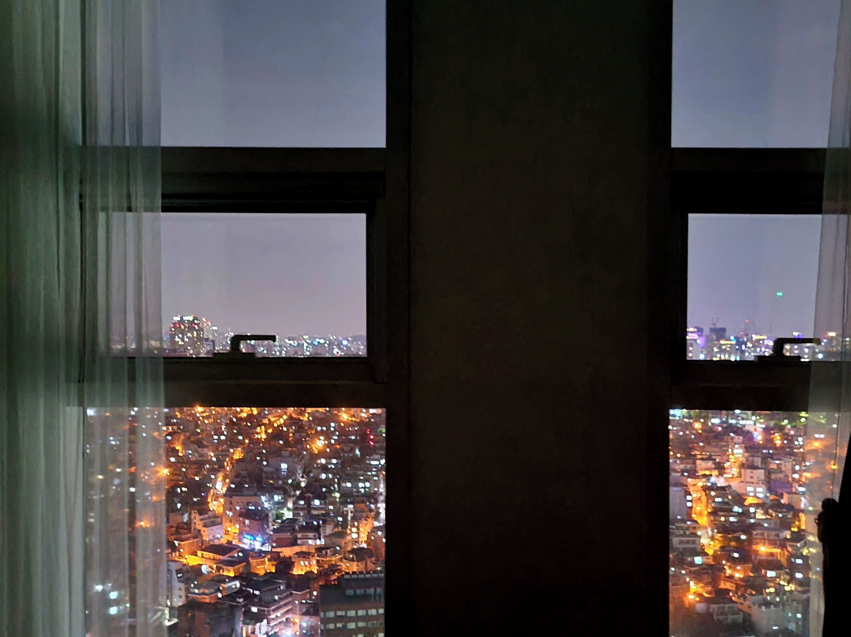 A view through a window with curtains, displaying a cityscape at night with illuminated buildings.