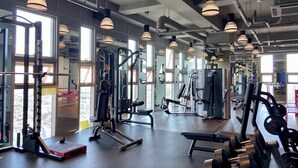 Un gimnasio con equipo de ejercicio diverso, ventanas amplias y lámparas colgantes.