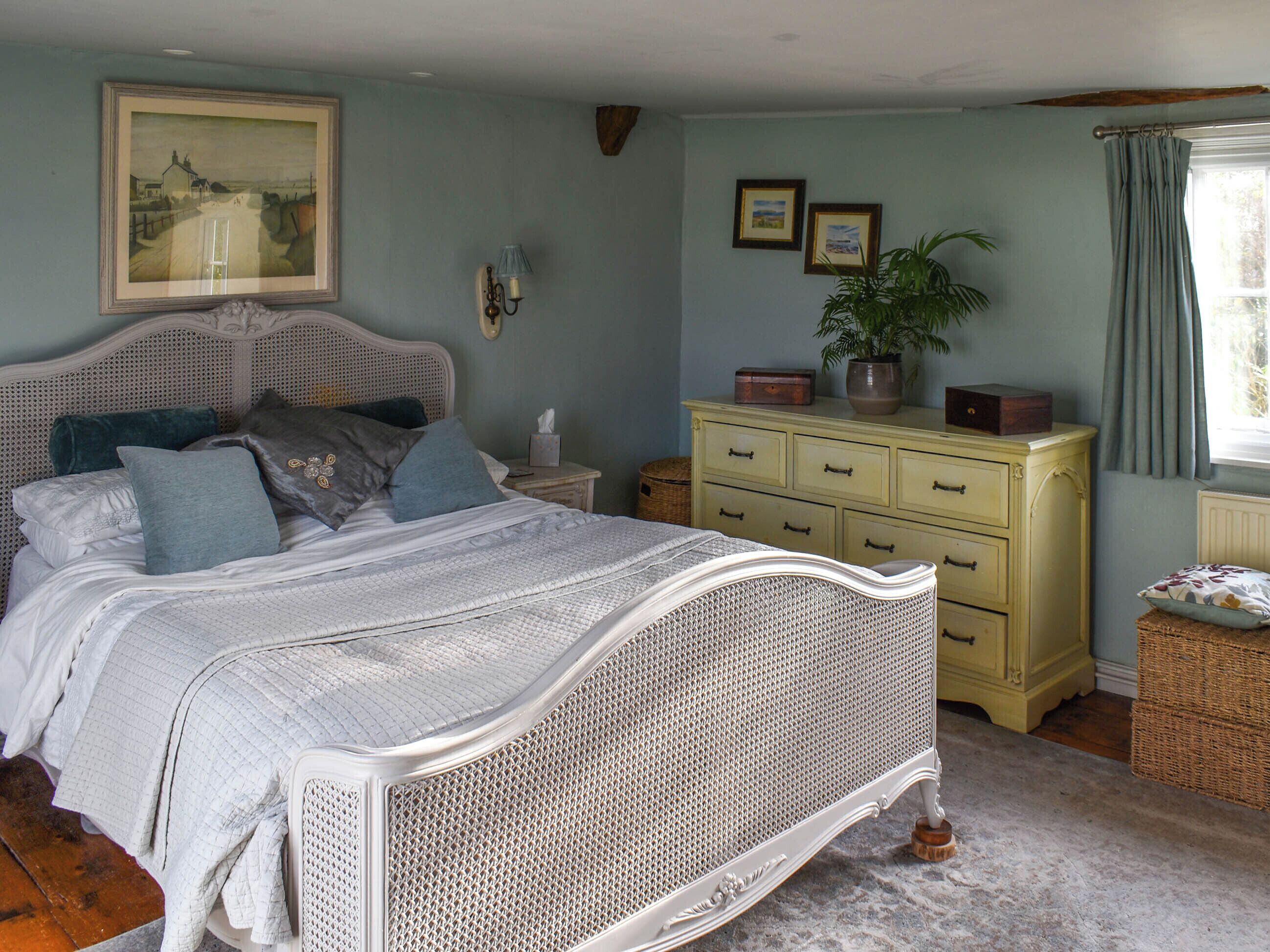 A bedroom with a white metal bed frame, a wooden nightstand, a yellow dresser, and a framed landscape painting on the wall.