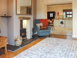 Un salon avec une cheminée, un fauteuil bleu, un tapis et, au fond, une cuisine.