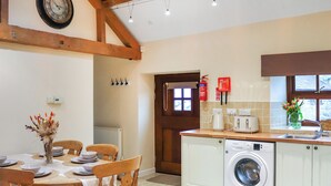 A kitchen with a washing machine, a dining table with chairs, a clock, and a fire extinguisher. - Beudy Newydd - UK50959 (Llanllechid, near Bangor)