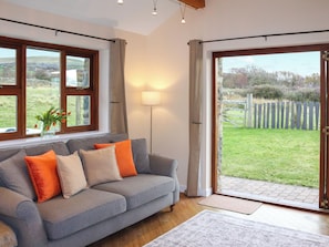 A living room with a grey sofa, orange and beige cushions, a floor lamp, and a view of a garden through a glass door. - Beudy Newydd - UK50959 (Llanllechid, near Bangor)