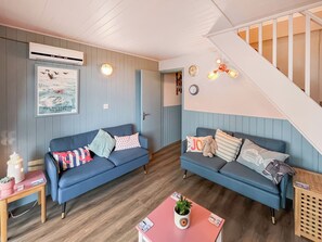 Un salon chaleureux avec deux canapés bleus, une table basse en bois, une petite table d’appoint et un escalier menant à l’étage.