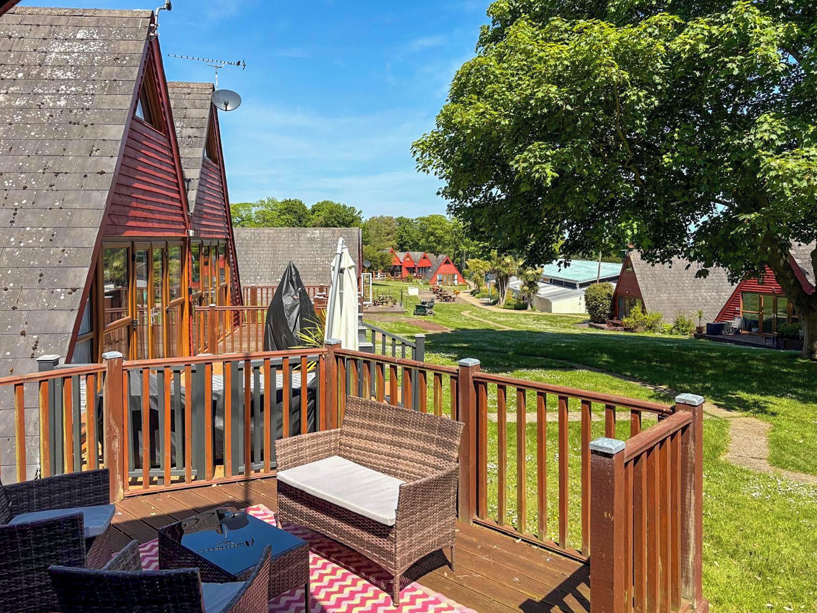 A deck with wicker furniture and a glass table, overlooking a grassy area with trees and buildings.