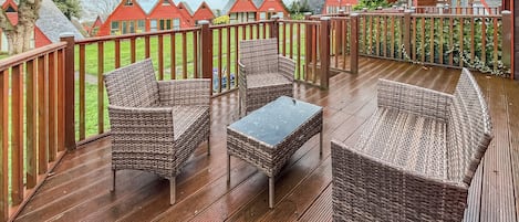 Una terraza de madera con mobiliario de mimbre y una mesa de vidrio, rodeada por una valla de madera.