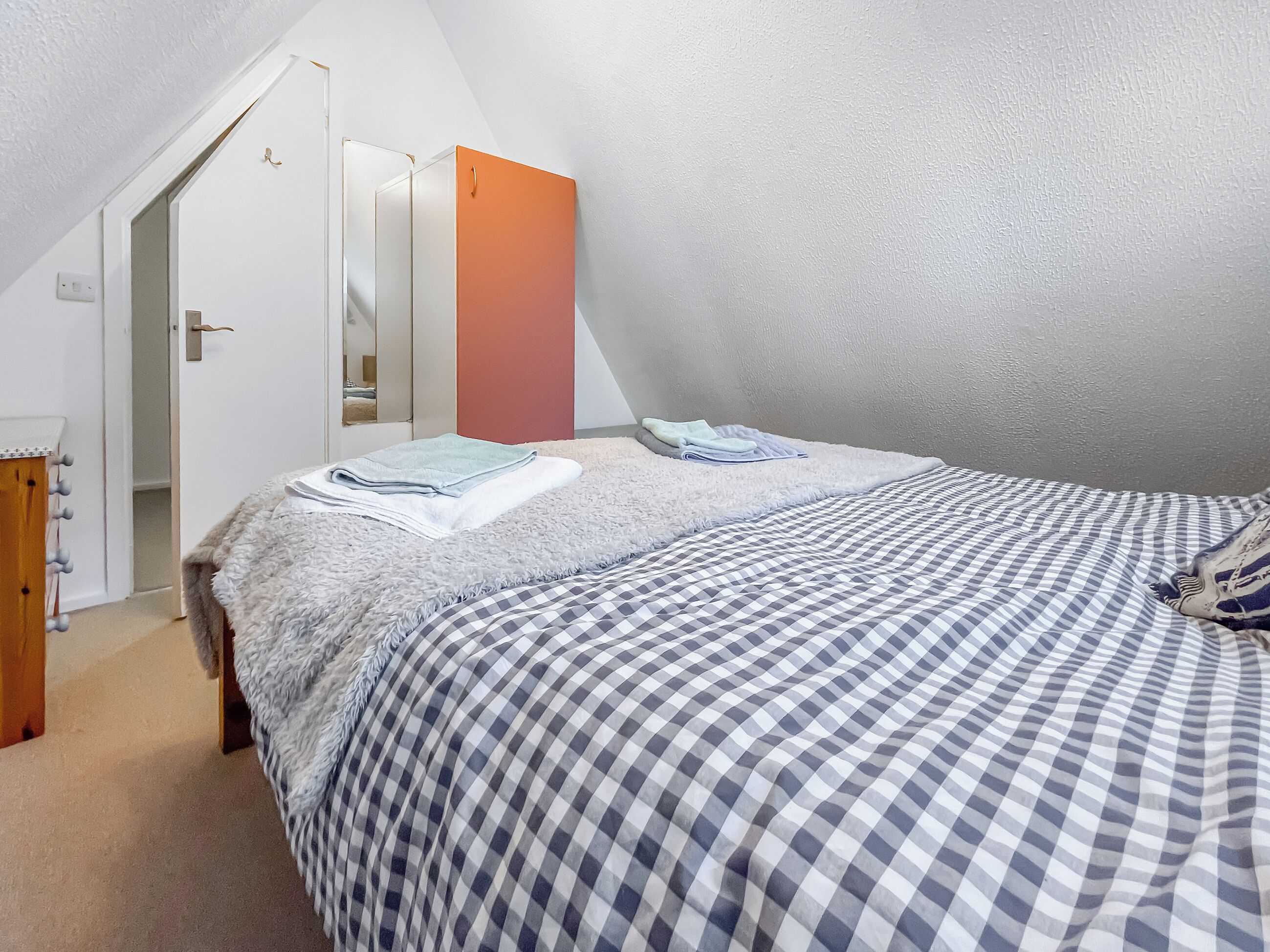 A bedroom with a checkered bedspread, a fluffy white blanket, and a folded towel. There is a door leading to a hallway with a mirror and a red cabinet.