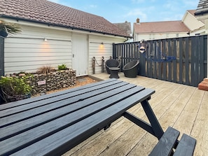 Eine Holzterrasse mit Picknicktisch, zwei Stühlen, einer Steinmauer mit Bepflanzung und einem Zaun.