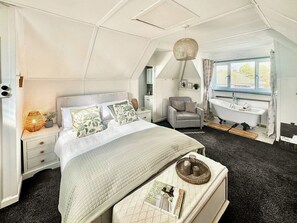 Et soveværelse med seng, sengebord, lampe, stol og et badeværelse med badekar og vindue.