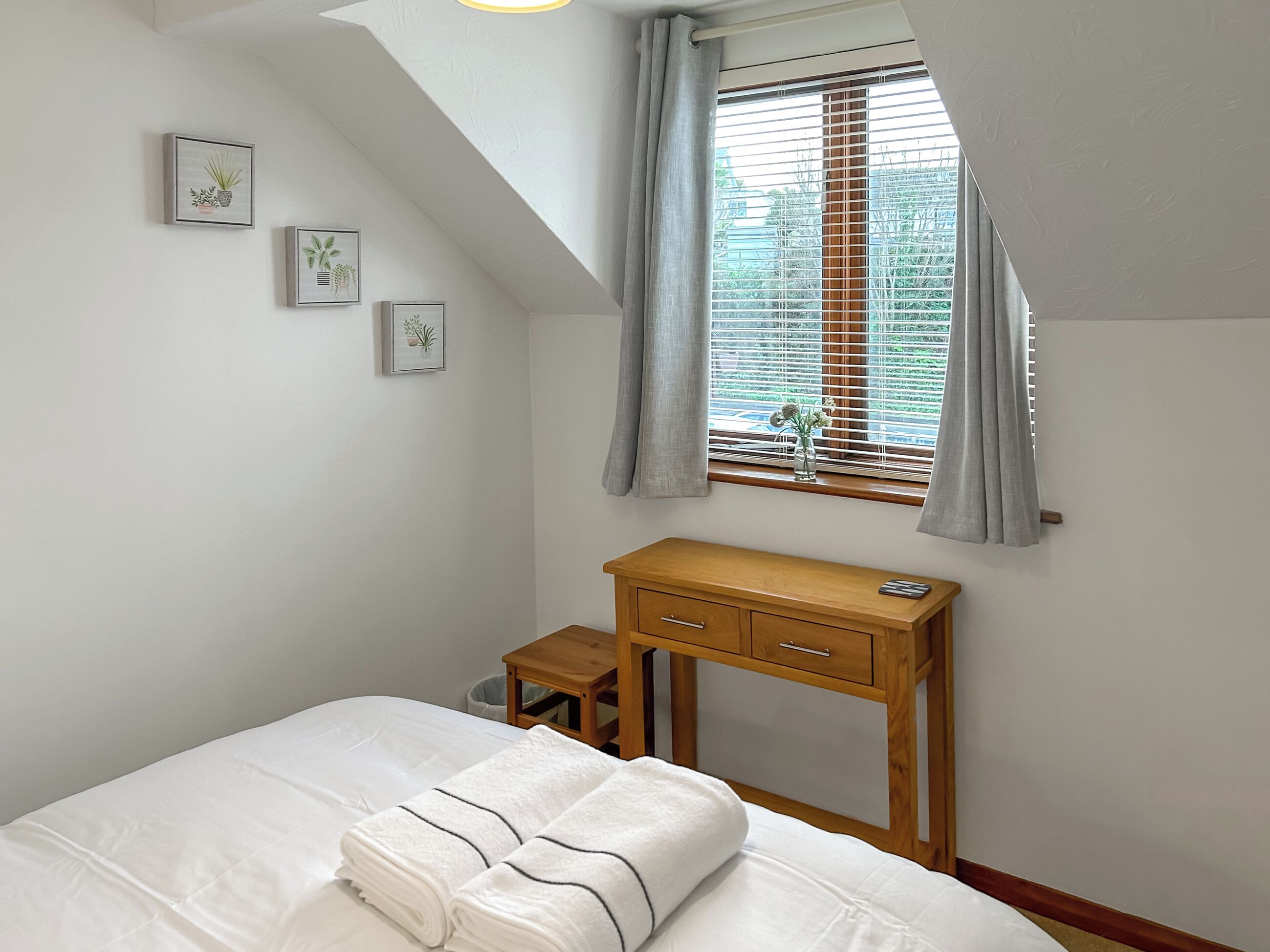 A bedroom with a bed, a wooden nightstand, a desk, and a window with blinds.