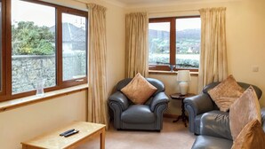 A living room with a sofa, armchair, and a small table. There are windows with curtains, a lamp on a side table, and a remote control on the table. - 4 bedroom accommodation in Brora (Brora)