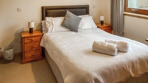 A neatly made bed with pillows, a wooden nightstand with a lamp, and a window with curtains. - 4 bedroom accommodation in Brora (Brora)