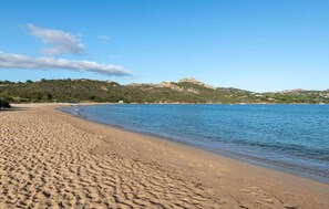 Ein Strand mit klarem, blauem Wasser, sandigem Ufer und einem Berg im Hintergrund.