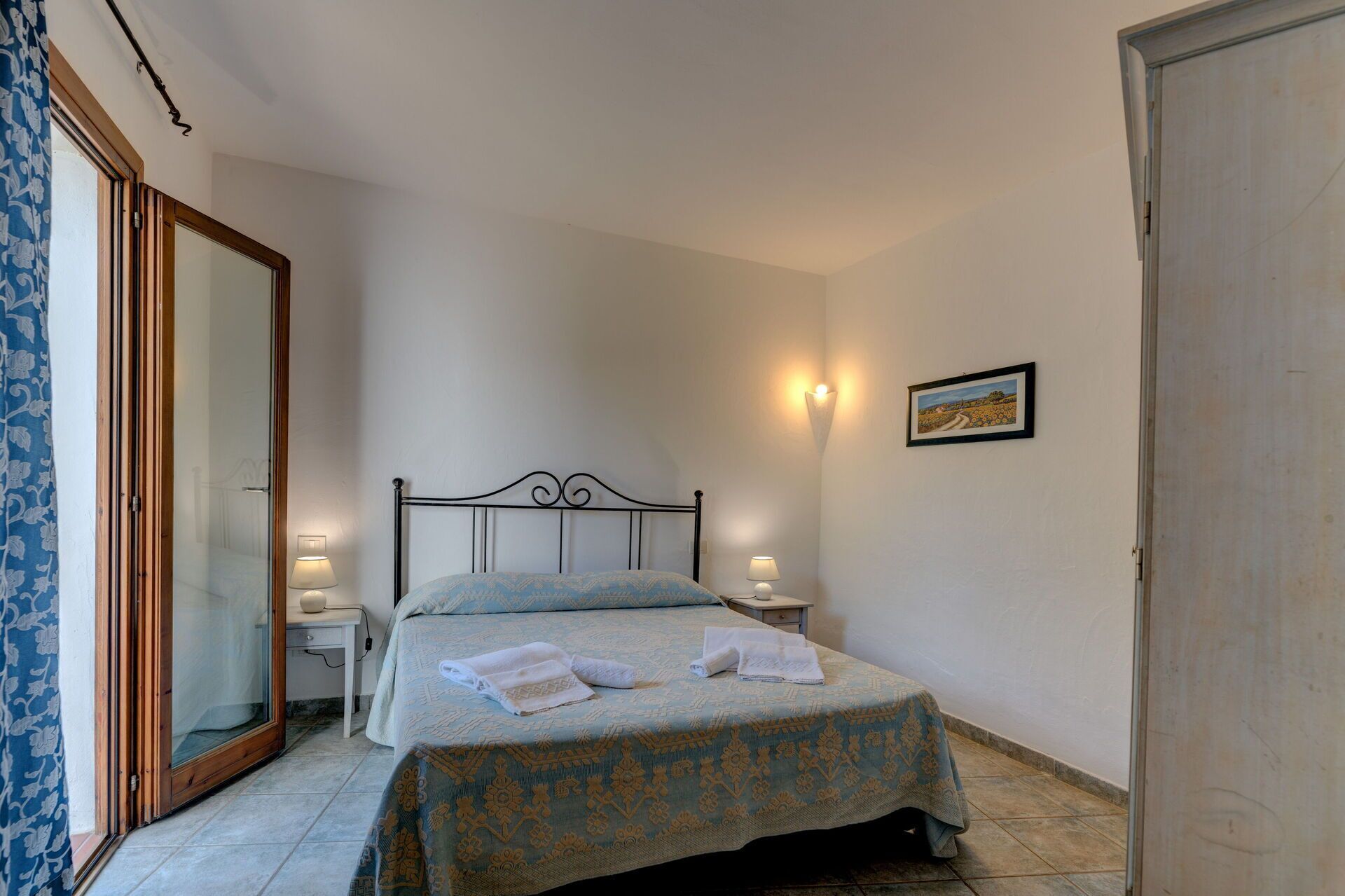 Une chambre à coucher avec un lit, deux tables de chevet avec des lampes, une porte et un tableau accroché au mur.