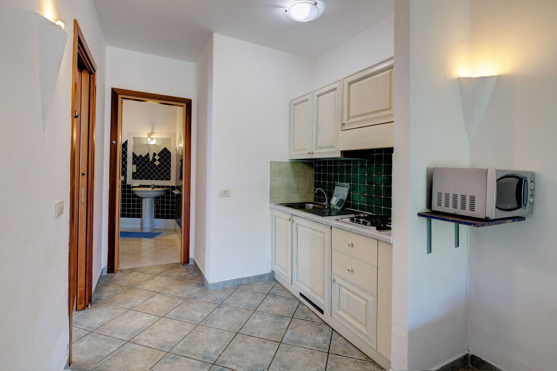 Un couloir étroit mène à une salle de bain équipée d’un lavabo et d’un miroir, à une cuisine avec des placards et un four à micro-ondes, et le sol est carrelé.