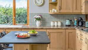 Een keuken met houten kastjes, een zwart aanrecht, een klok en een raam met uitzicht op groen.