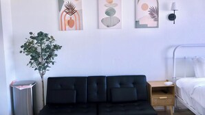 Habitación de hotel moderna con un sofá negro, una mesita de noche de madera, una mesita auxiliar y una cama. Hay pinturas abstractas en la pared y una planta en maceta.