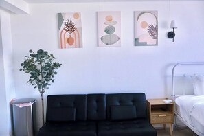 Ein modernes Hotelzimmer mit einer schwarzen Couch, einem hölzernen Nachttisch, einem Beistelltisch und einem Bett. An der Wand befinden sich abstrakte Gemälde und ein Blumentopf.