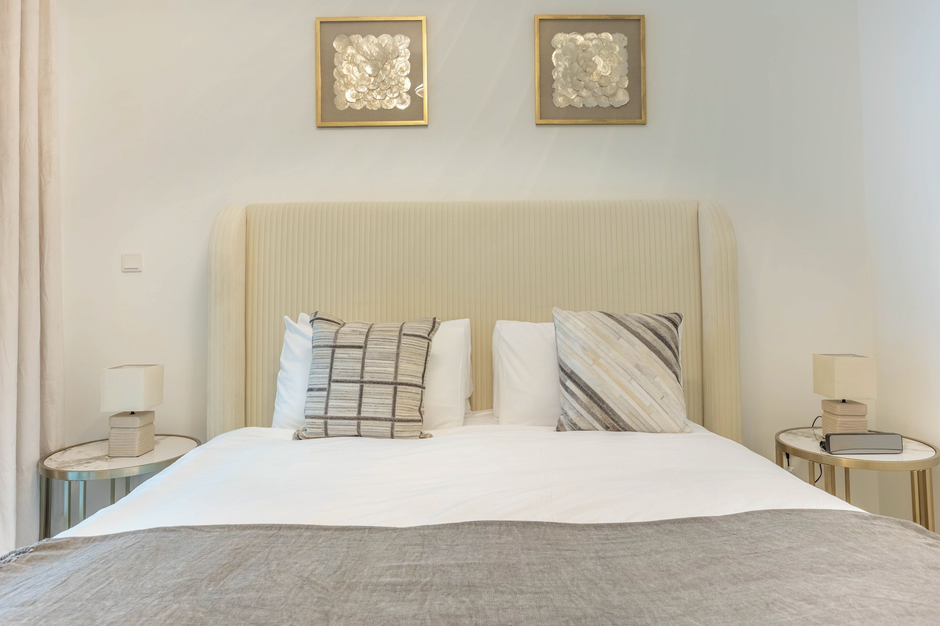 Un lit bien fait, avec une couette blanche et deux oreillers, flanqué de deux tables de chevet sur lesquelles reposent des lampes, dans une pièce dont le mur est uni et qui est décorée de deux œuvres d’art encadrées.
