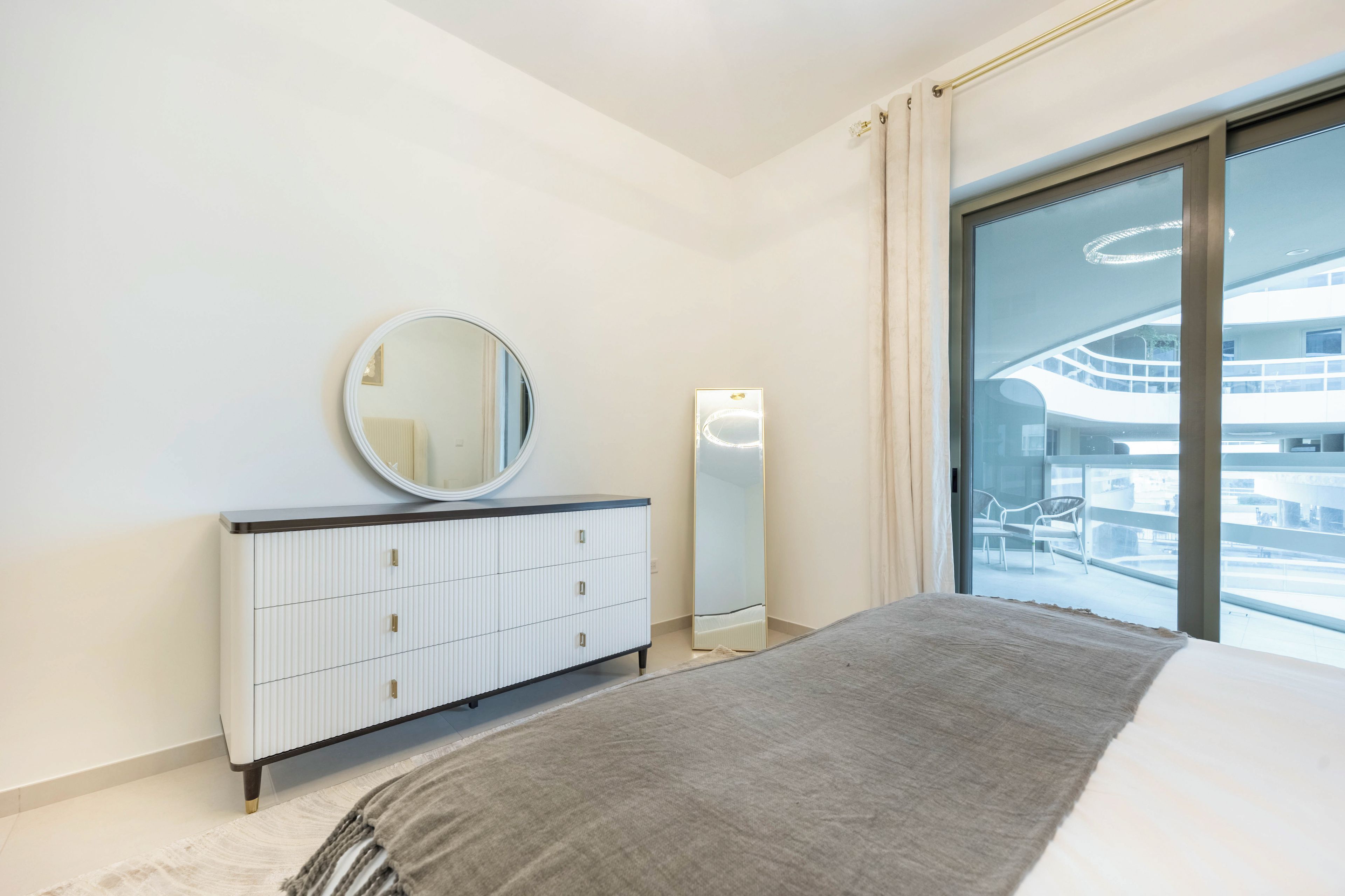 A modern bedroom with a bed, a dresser, a mirror, and a large window with a view of a building.