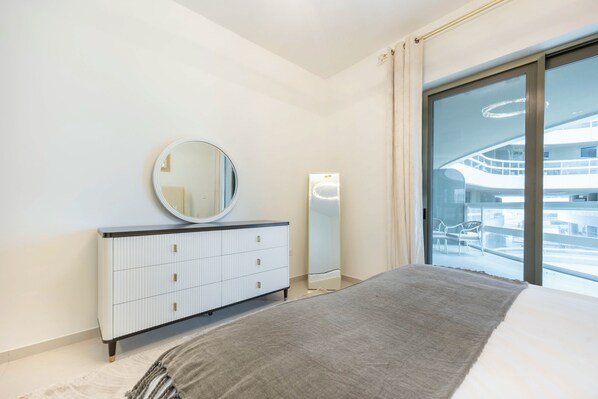 A modern bedroom with a bed, a dresser, a mirror, and a large window with a view of a building.