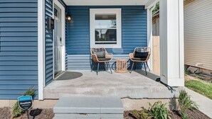 Une maison avec un porche abrité, deux fauteuils en osier, une petite table et un panneau indiquant « Zone protégée par SimpliSafe ».